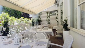 Une terrasse extérieure avec des tables et des chaises blanches, protégée par un auvent et entourée de verdure et de plantes.