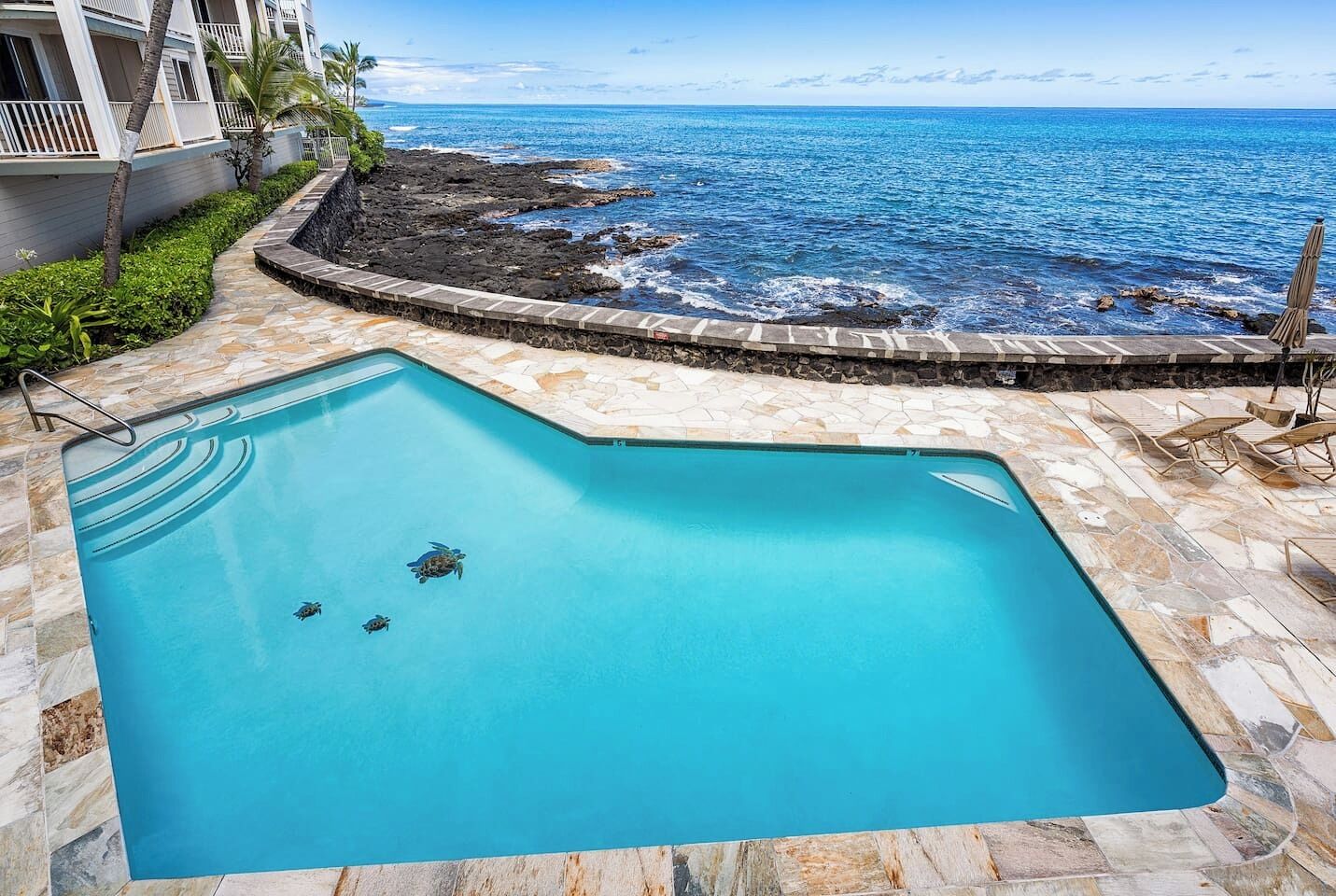 A swimming pool with a clear view of the ocean and a stone pathway leading to a building with balconies.