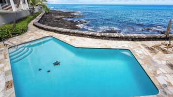 A swimming pool with a clear view of the ocean and a stone pathway leading to a building with balconies.
