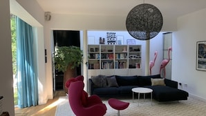 Ein modernes Wohnzimmer mit einem dunkelgrauen Sofa, roten Sesseln, einem weißen Couchtisch, einer großen Topfpflanze und einem Bücherregal voller Bücher.