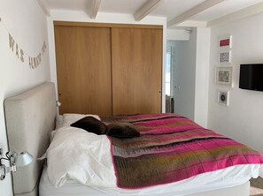 A bedroom with a bed, a wooden wardrobe, a wall-mounted TV, and a door leading to another room.