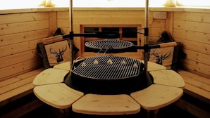 Hyggelig træbeklædt interiør med et centralt grillbord omgivet af bænke, en sort skorsten og et vindue med naturligt lys.