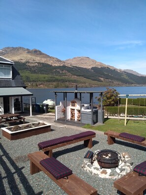 A patio area with wooden benches, a fire pit, and a barbecue grill, overlooking a lake with mountains in the background.