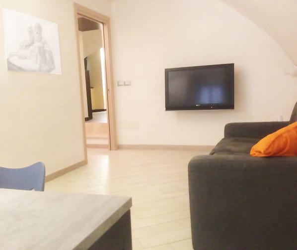 A living room with a flat-screen TV mounted on the wall, a dark-colored sofa with an orange cushion, and a wooden dining table.