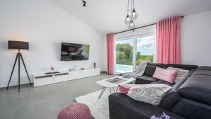 Ett modernt vardagsrum med en stor platt-TV, en mörk soffa, ett vitt soffbord och en rosa pall.