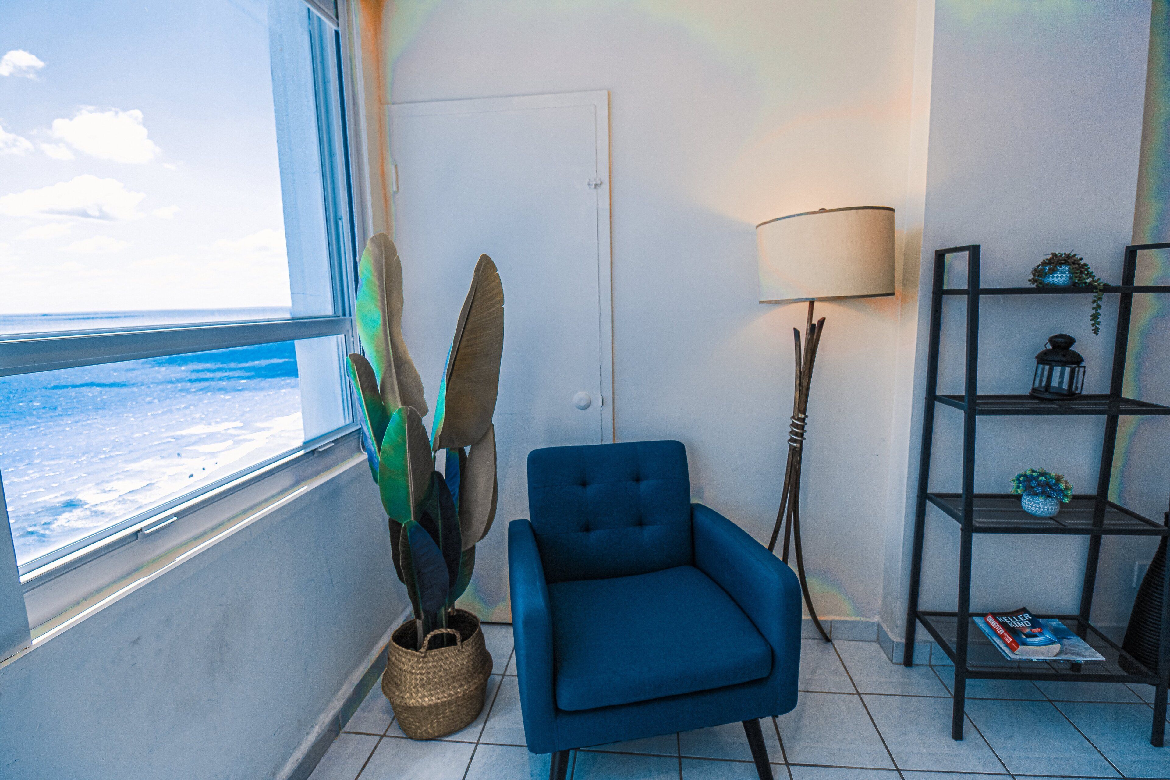 A room with a view of the ocean, a blue armchair, a lamp, a bookshelf, and a potted plant.