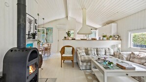 A cozy living room with a wood-burning stove, a dining area with a table and chairs, a sofa, and a large window. - 4 bedroom amazing home in Hornbæk (Hornbæk)