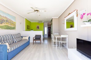 Una sala de estar con un sofá azul, una televisión, un ventilador de techo y un comedor con una mesa y sillas blancas.