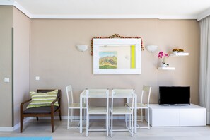 Un salón moderno con pared beige, un cuadro enmarcado, estanterías blancas con flores, un televisor y una mesa de comedor con sillas.