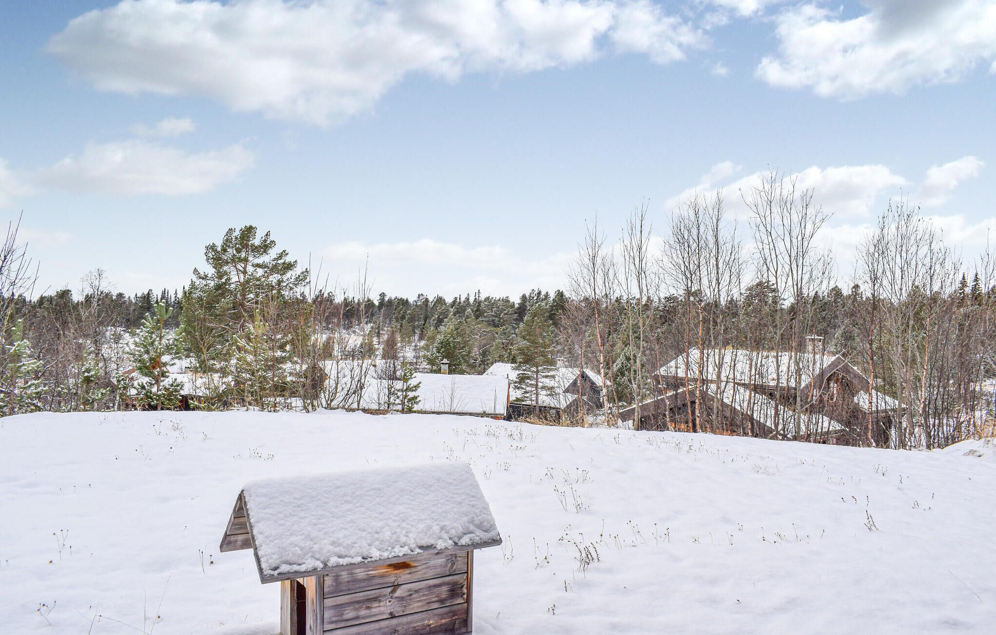 A wooden cabin with a snow-covered roof in a snowy landscape with houses and trees in the background.