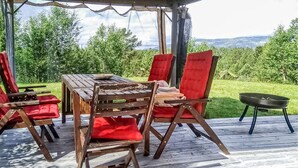 A covered outdoor dining area with a wooden table and red cushioned chairs, overlooking a grassy landscape with trees and hills.