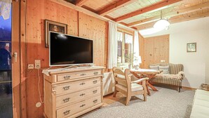 L’intérieur d’un chalet en bois chaleureux et accueillant, avec une télévision posée sur une commode, une table à manger avec des chaises, un canapé et une fenêtre avec des rideaux.