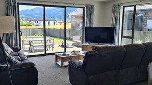 Een woonkamer met een bank, een salontafel en een televisie. Er is een glazen schuifdeur die leidt naar een buitenruimte met een tafel en stoelen.