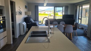 Een moderne keuken met een centraal eiland, een eethoek met een tafel en stoelen, en een grote schuifpui die leidt naar een balkon met uitzicht op de omgeving.