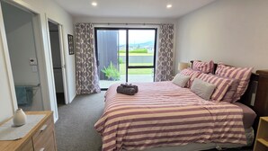 Een slaapkamer met een bed, nachtkastje, lamp, raam met gordijnen en uitzicht op het omliggende landschap.