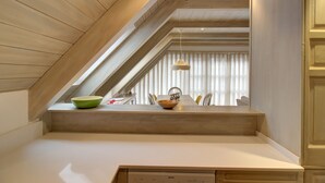 Un espace repas doté d’un plafond haut, de poutres en bois et offrant une vue sur la pièce grâce à une fenêtre munie de stores.