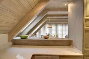 Zona pranzo con soffitto alto, travi in legno e vista sulla stanza attraverso una finestra con persiane.