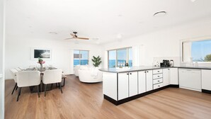 A modern kitchen with white cabinetry, a dining area with a white table and chairs, and a view of the sea through large windows.