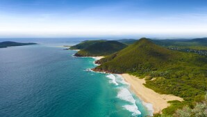 An aerial view of a coastal area with a beach, clear blue waters, and green hills.
