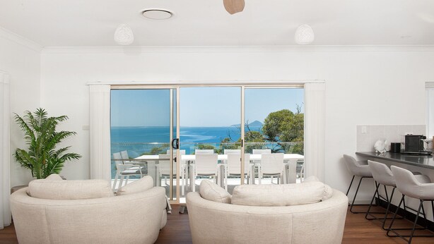 A modern living room with a large sliding glass door leading to a balcony with a view of the sea. There are two white sofas and a dining table with chairs.