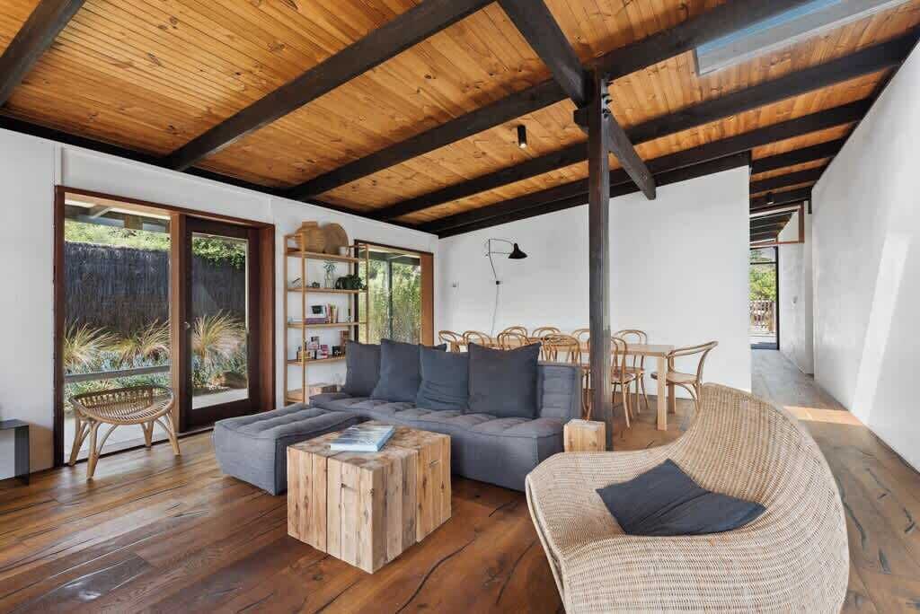 A modern living room with a wooden ceiling, a grey sofa, wicker furniture, and a view through glass doors.