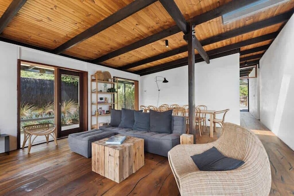 A modern living room with a wooden ceiling, a grey sofa, wicker furniture, and a view through glass doors.
