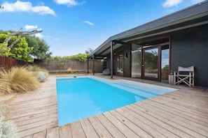 A swimming pool with a wooden deck, a covered patio area, and a view of a natural landscape.