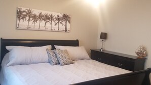 Un lit bien fait, avec une literie blanche et des oreillers à motifs, une tête de lit sombre, une table de chevet fixée au mur avec une lampe, et une maquette de bateau posée sur la table de chevet.