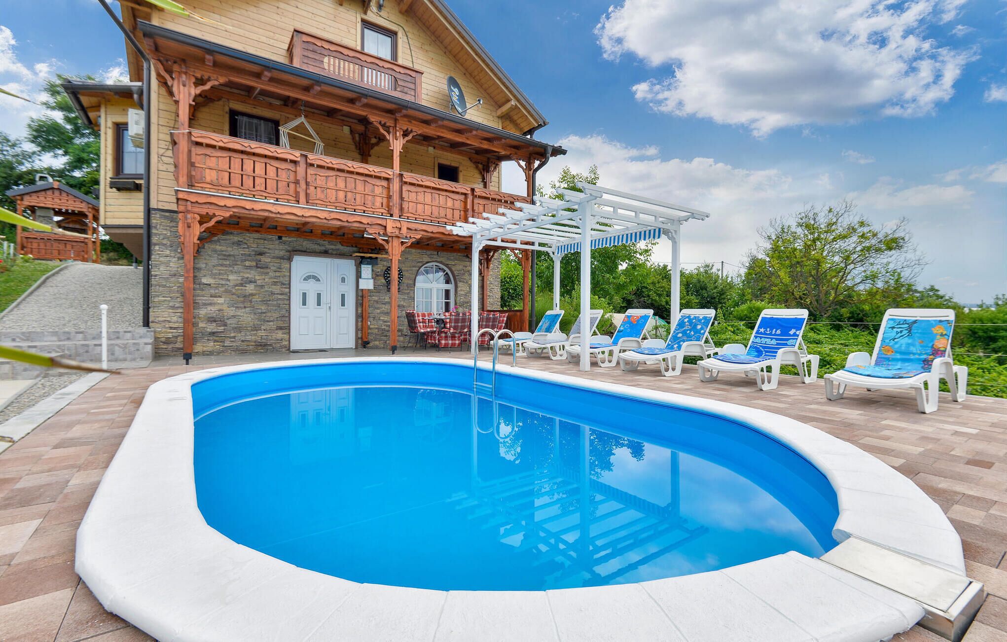 Una piscina con sillas de descanso, una casa de madera con balcón y un muro de piedra.