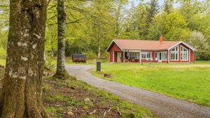 A red wooden cabin with a gravel path leading to it, surrounded by lush greenery and trees.