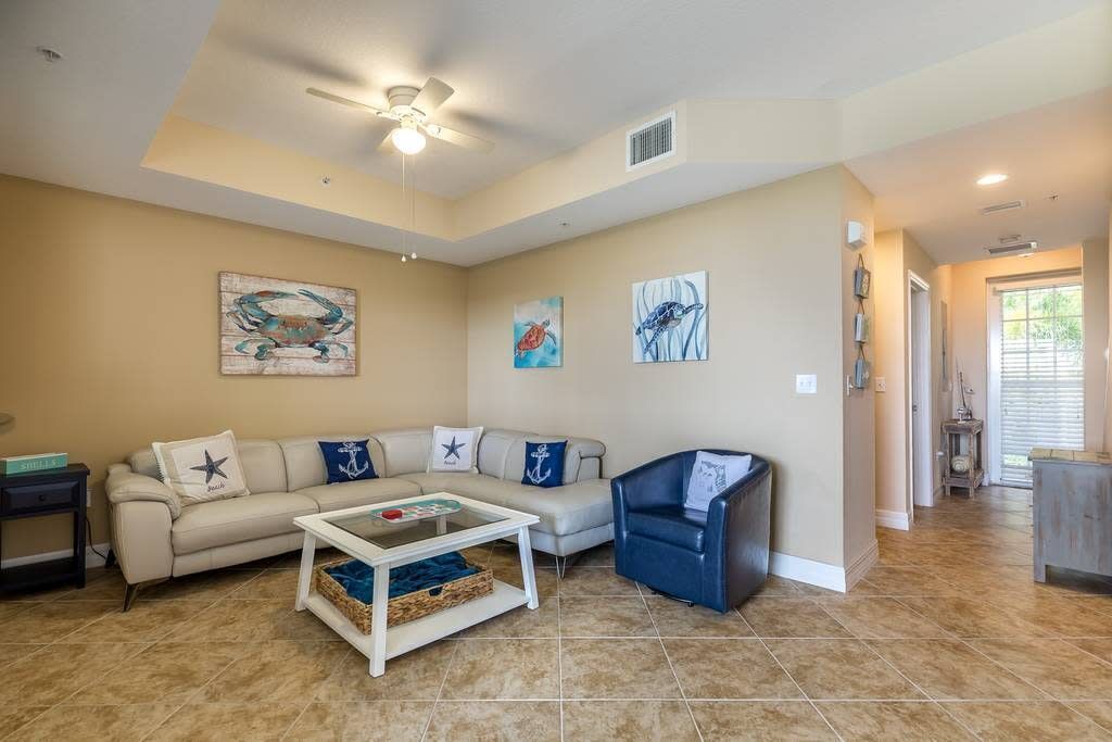 A living room with a beige sofa, a coffee table, and a blue armchair. There are three paintings on the wall and a ceiling fan.