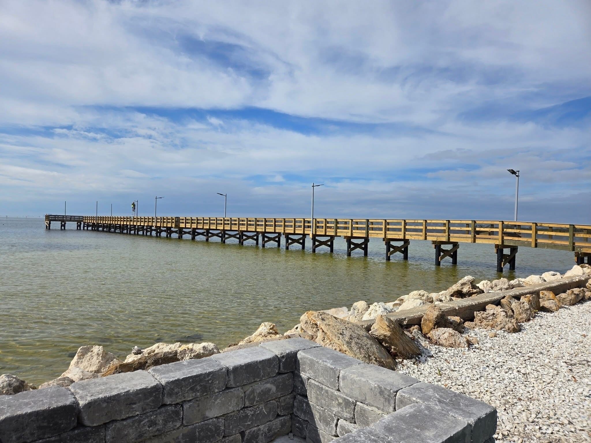 A long wooden pier extending into calm waters, supported by sturdy pillars, with a stone wall in the foreground.