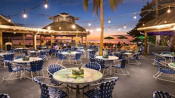 Un espace de restauration en plein air avec des tables rondes et des chaises bleues, entouré de guirlandes lumineuses et de palmiers.