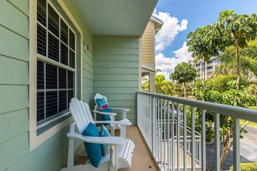 A balcony with two white Adirondack chairs and a small table, overlooking a residential area with houses and greenery.