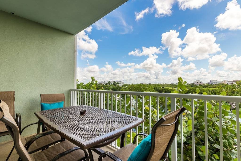 A balcony with a table and chairs, a view of greenery and buildings, and a clear sky with clouds.