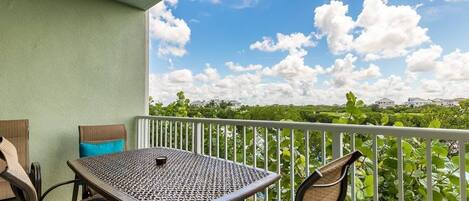 A balcony with a table and chairs, a view of greenery and buildings, and a clear sky with clouds.
