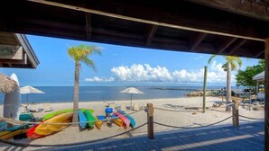 Une terrasse en bois donnant sur une plage, avec des kayaks aux couleurs vives, des parasols et des chaises longues.