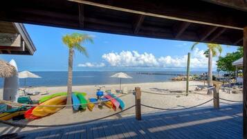 Une terrasse en bois donnant sur une plage, avec des kayaks aux couleurs vives, des parasols et des chaises longues.