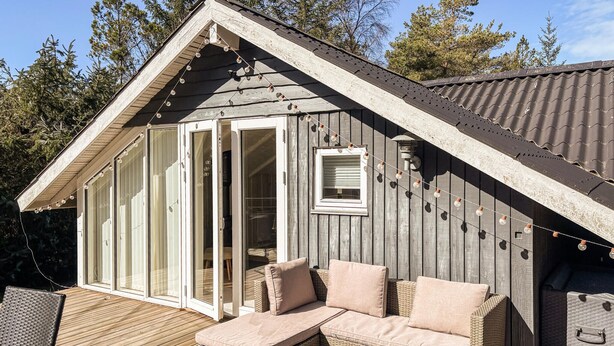 A wooden cabin with a sloped roof, large glass sliding doors, a wicker sofa with cushions, and a small table with a plant. - Amazing home in ålbæk with WiFi (Ålbæk)