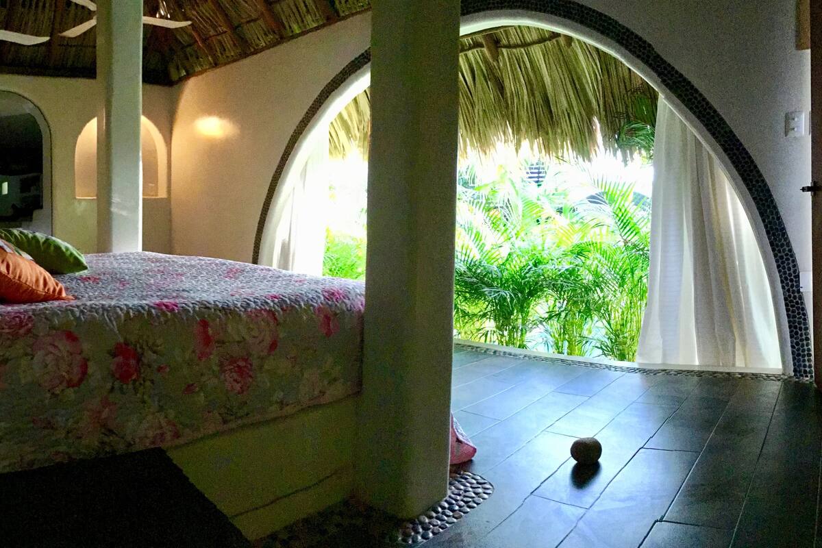 A bed with floral bedding and pillows, a thatched ceiling, a view of greenery through an arched window, and a tiled floor.