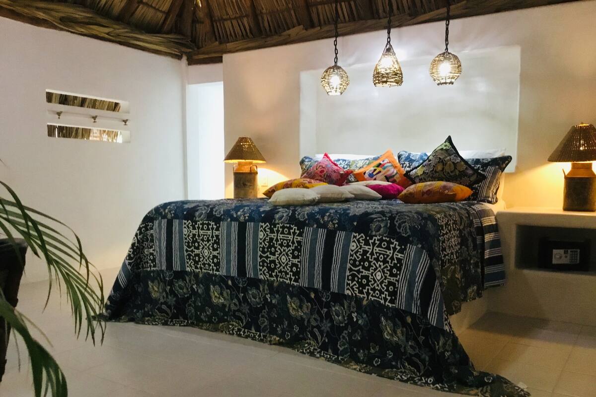 A bedroom with a patterned bedspread, multiple pillows, hanging pendant lights, and a small bedside table with a lamp.
