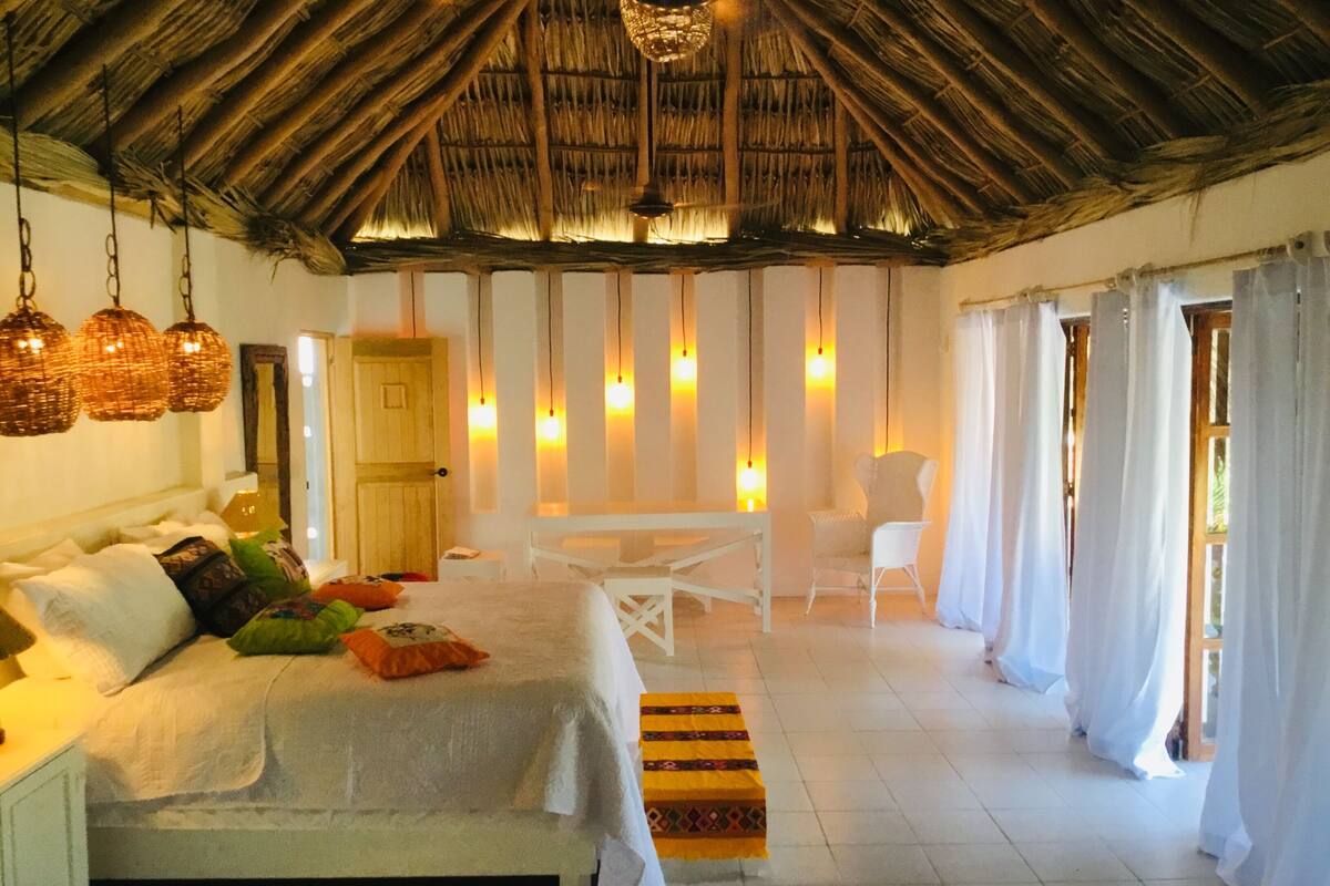 A spacious bedroom with a large bed, a wooden ceiling, hanging lights, a small table, and a chair.