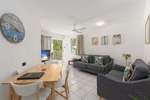 A modern living room with a sofa, a dining table with chairs, a large wall clock, and a ceiling fan. - Seascape Holidays at The Queenslander One Bedroom (Douglas)