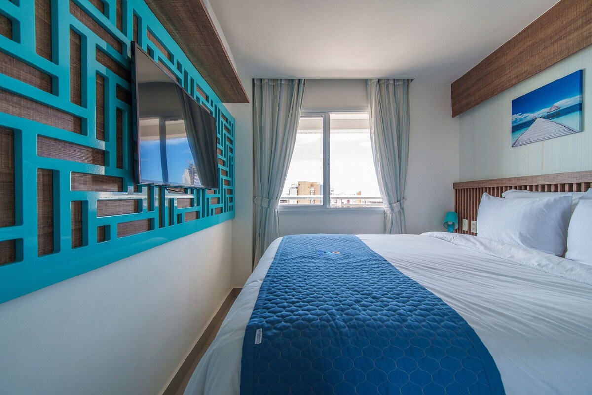 A hotel room with a bed, a TV, a window with curtains, and a painting on the wall.