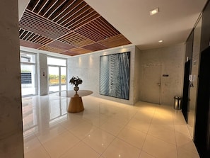 A modern lobby with a wooden ceiling, a large abstract painting, a round table with a vase, and a door with a trash bin. - Compacto de Luxo - Helbor My Way (Fortaleza)