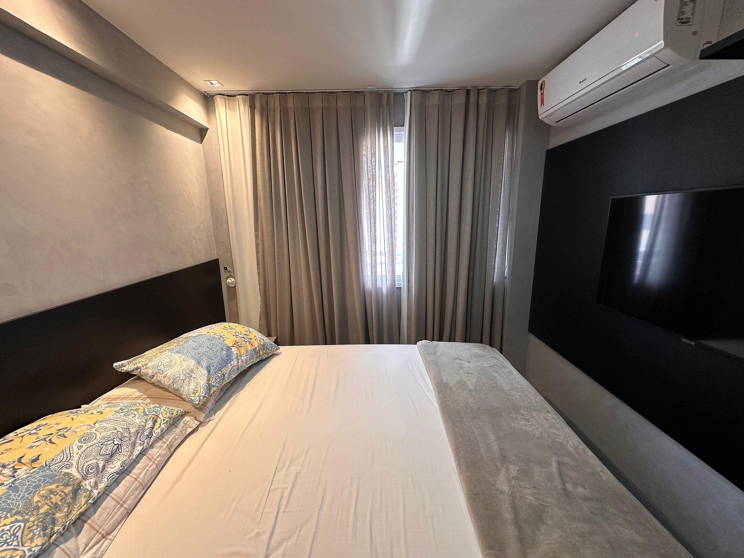 A hotel room with a bed, a TV, an air conditioner, and a window with curtains.