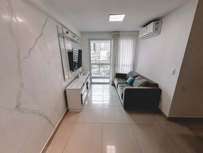 A modern living room with a flat-screen TV, a sofa, a coffee table, and a sliding glass door leading to a balcony. - Compacto de Luxo - Helbor My Way (Fortaleza)