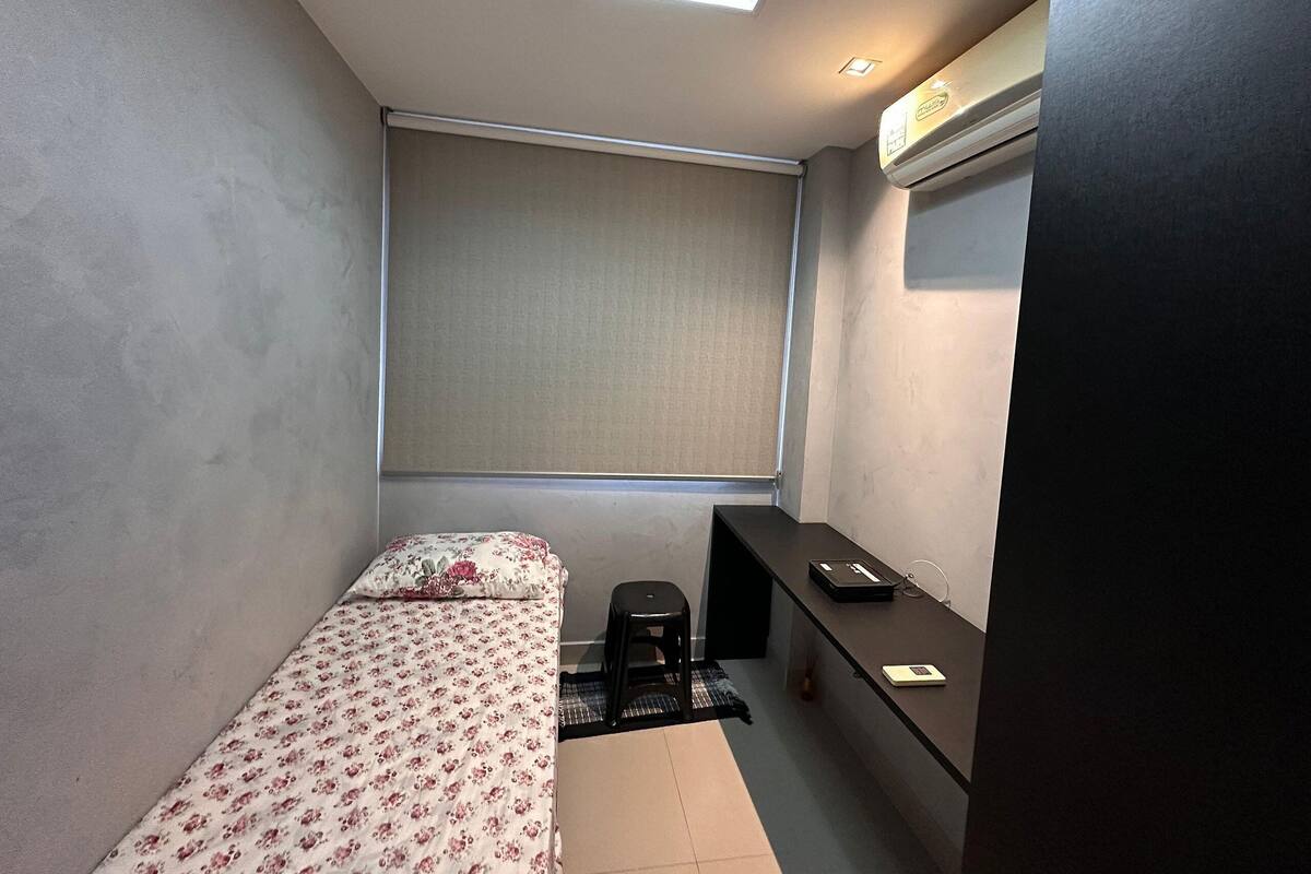 A small, compact room with a single bed, a desk, a chair, a window with a blind, and a ceiling light.