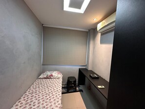 A small, compact room with a single bed, a desk, a chair, a window with a blind, and a ceiling light. - Compacto de Luxo - Helbor My Way (Fortaleza)