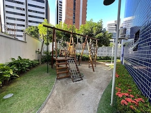Ein Spielplatz mit Holzkonstruktionen und einer Rutsche, umgeben von einem kleinen Garten und einer blauen Fliesenwand.